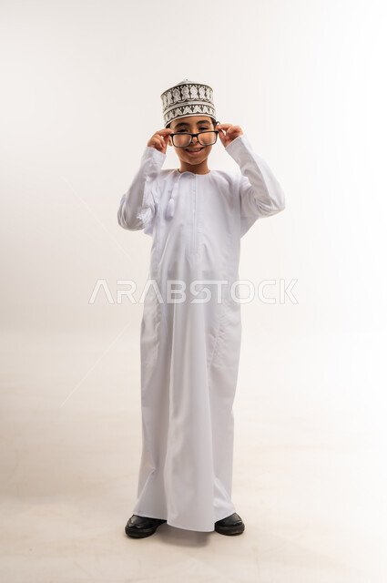 النظر إلى الكاميرا بإيماءات السرور والسعادة، الاهتمام بالمظهر الخارجي, بورتريه لولد عربي خليجي عماني يرتدي الدشداشة و الكمة يقوم بارتداء النظارات الطبية، مفهوم الاناقة والوسامة، صورة الجسم بالطول الكامل, خلفية بيضاء