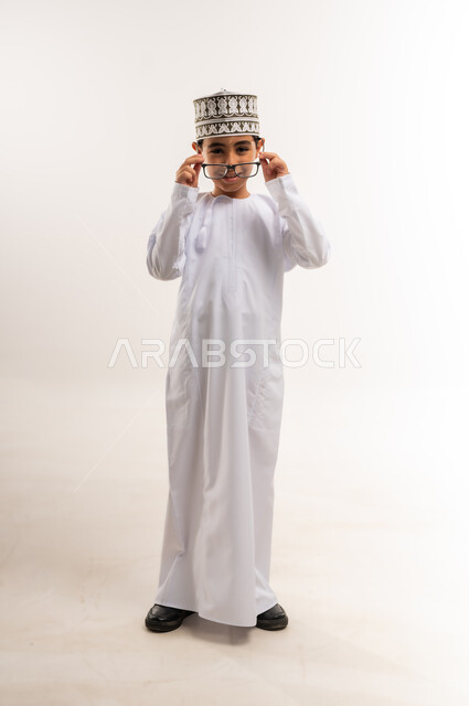 الاهتمام بالمظهر الخارجي, بورتريه لولد عربي خليجي عماني يرتدي الدشداشة و الكمة يقوم بارتداء النظارات الطبية ينظر إلى الكاميرا بإيماءات السرور والسعادة، مفهوم الاناقة والوسامة، صورة الجسم بالطول الكامل, خلفية بيضاء