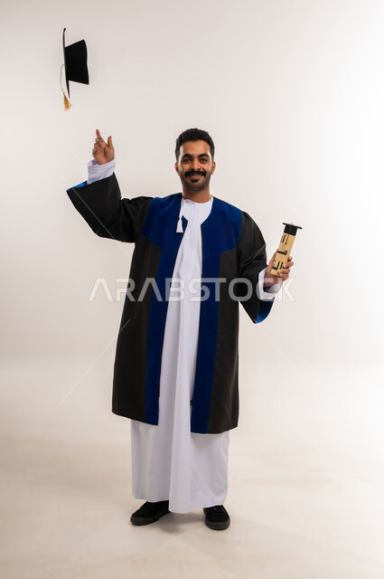 السعادة بالوصول لحلم النجاح، بورتريه لخريج عربي خليجي عماني يرتدي الدشداشة وزي التخرج يمسك بيده وثيقة التخرج يقوم برمي القبعة للأعلى ينظر إلى الكاميرا بإيماءات السرور، مفهوم الاجتهاد والتفوق، الاحتفال بإنهاء الدراسة الجامعية، الجسم بالطول الكامل، خلفية بيضاء