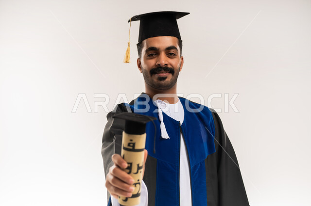 الاحتفال بإنهاء الدراسة الجامعية، إيماءات السعادة بالوصول لحلم النجاح، بورتريه مقرب لخريج عربي خليجي عماني يرتدي الدشداشة وزي التخرج والقبعة يمسك بيده وثيقة التخرج ينظر إلى الكاميرا بتعابير الرضا والسرور، مفهوم الاجتهاد والتفوق، خلفية بيضاء