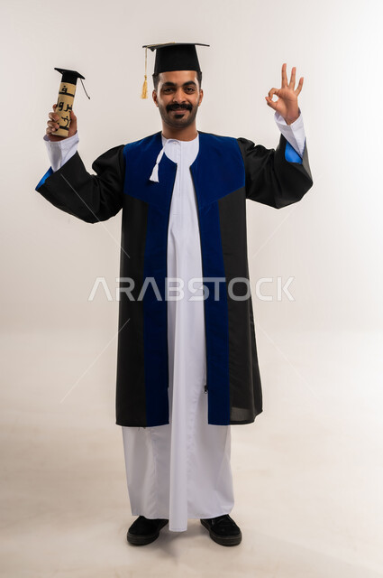 علامة الجودة والامتياز، السعادة بالوصول لحلم النجاح، بورتريه لخريج عربي خليجي عماني يرتدي الدشداشة وزي التخرج يمسك وثيقة التخرج يرفع يده ينظر إلى الكاميرا بتعابير الإعجاب، مفهوم الاجتهاد والتفوق، الاحتفال بإنهاء الدراسة الجامعية، الجسم بالطول الكامل، خلفية بيضاء