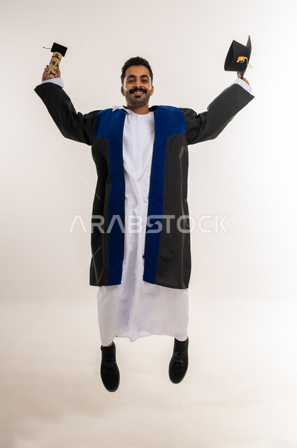 القفز عالياً بتعابير الفرح والحماس، الاحتفال بإنهاء الدراسة الجامعية، السعادة بالوصول لحلم النجاح، بورتريه لخريج عربي خليجي عماني يرتدي الدشداشة وزي التخرج يمسك القبعة ووثيقة التخرج ينظر إلى الكاميرا، مفهوم الاجتهاد والتفوق، الجسم بالطول الكامل، خلفية بيضاء