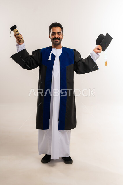 الاحتفال بإنهاء الدراسة الجامعية، السعادة بالوصول لحلم النجاح، بورتريه لخريج عربي خليجي عماني يرتدي الدشداشة وزي التخرج يمسك القبعة ووثيقة التخرج ينظر إلى الكاميرا بتعابير السرور والرضا، مفهوم الاجتهاد والتفوق، الجسم بالطول الكامل، خلفية بيضاء