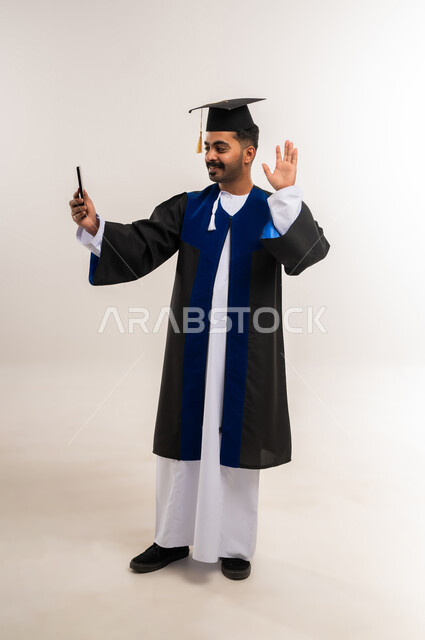 مشاركة الأهل والأصدقاء فرحة الوصول لحلم النجاح، استخدام جهاز تقني حديث، بورتريه لخريج عربي خليجي عماني يرتدي الدشداشة وزي التخرج والقبعة يرفع يده يقوم بإجراء مكالمة فيديو عبر الهاتف المحمول، إلقاء التحية والسلام، الجسم بالطول الكامل، خلفية بيضاء