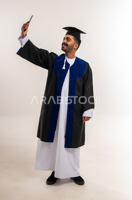 التقاط صورة سيلفي تذكارية عبر الجوال، استخدام جهاز تقني حديث، إيماءات السعادة بالوصول لحلم النجاح، بورتريه لخريج عربي خليجي عماني يرتدي الدشداشة وزي التخرج والقبعة يمسك بيده الهاتف المحمول، مفهوم التفوق والاجتهاد، الجسم بالطول الكامل، خلفية بيضاء