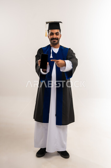 Showing a blank black screen, using a modern technical device, gestures of happiness at reaching the dream of success, portrait of an Arab Gulf Omani graduate wearing a dishdasha, graduation uniform and a hat, holding a mobile phone in his hand and pointing at it, the concept of excellence and diligence, full-length body, white background