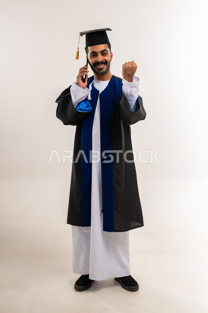استخدام جهاز تقني حديث، مشاركة الأهل والأصدقاء فرحة الوصول لحلم النجاح، بورتريه لخريج عربي خليجي عماني يرتدي الدشداشة وزي التخرج والقبعة يقوم بإجراء مكالمة هاتفية عبر الهاتف المحمول يرفع قبضة يده بإيماءات الفوز والنصر، الجسم بالطول الكامل، خلفية بيضاء