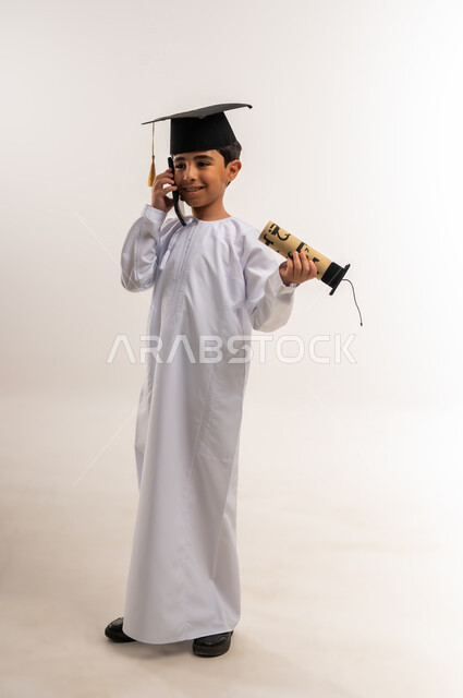 الاحتفال بالتخرج من الفصل الدراسي، مشاركة الأهل والأصدقاء فرحة تحقيق النجاح، استخدام جهاز تقني حديث، بورتريه لطالب مدرسي عربي خليجي عماني يرتدي الدشداشة وقبعة التخرج يمسك وثيقة التخرج يقوم بإجراء مكالمة هاتفية عبر الهاتف المحمول، الجسم بالطول الكامل، خلفية بيضاء