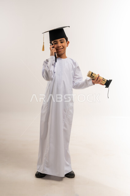 مشاركة الأهل والأصدقاء فرحة تحقيق النجاح، استخدام جهاز تقني حديث، بورتريه لطالب مدرسي عربي خليجي عماني يرتدي الدشداشة وقبعة التخرج يمسك وثيقة التخرج يقوم بإجراء مكالمة هاتفية عبر الهاتف المحمول، الاحتفال بالتخرج من الفصل الدراسي، الجسم بالطول الكامل، خلفية بيضاء