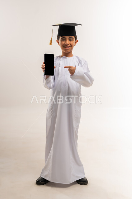 استخدام جهاز تقني حديث، تعابير الفخر والسعادة لتحقيق النجاح، بورتريه لطالب مدرسي عربي خليجي عماني يرتدي الدشداشة وقبعة التخرج يمسك بيده الهاتف المحمول يشير إليه، عرض شاشة سوداء فارغة، الاحتفال بالتخرج من الفصل الدراسي، الجسم بالطول الكامل، خلفية بيضاء