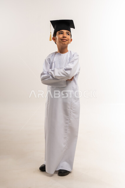 الانتقال إلى سنة دراسية جديدة، تعابير الفخر والسعادة لتحقيق النجاح، بورتريه لطالب مدرسي عربي خليجي عماني يرتدي الدشداشة وقبعة التخرج يقف بأيدي متقاطعة ينظر إلى الأعلى بإيماءات السرور، الاحتفال بالتخرج من الفصل الدراسي، الجسم بالطول الكامل، خلفية بيضاء