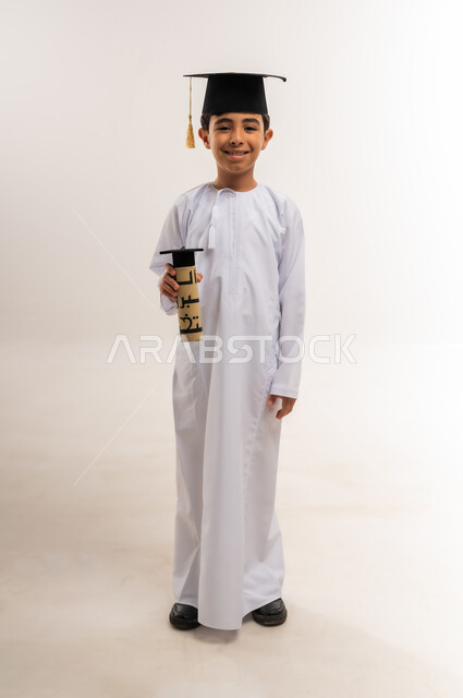 Celebrating graduation from the school term, expressions of pride and happiness for achieving success, portrait of an Arab Gulf Omani school student wearing a dishdasha and graduation cap holding his graduation document looking at the camera with gestures of pleasure, moving to a new school year, full body, white background