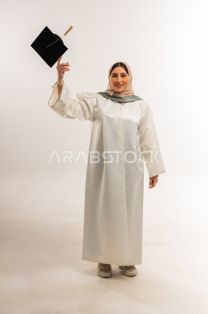 إيماءات السعادة بالوصول لحلم النجاح، بورتريه لخريجة عربية خليجية عمانية محجبة مبتسمة ترتدي زي التخرج تقوم برمي القبعة بتعابير السرور والحماس، شابة سعودية تحتفل بإنهاء الدراسة الجامعية، حفلة التخرج من الجامعة، الجسم بالطول الكامل، خلفية بيضاء