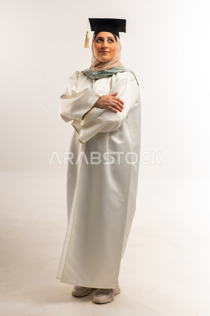 University graduation party, gestures of happiness at reaching the dream of success, portrait of a smiling veiled Gulf Arab Omani graduate wearing graduation dress and cap standing with crossed hands looking up with expressions of pleasure, young Saudi woman celebrating finishing university studies, full body, white background