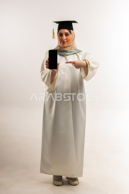 شابة سعودية تستخدم جهاز تقني حديث، عرض شاشة سوداء فارغة، إيماءات السعادة بالوصول لحلم النجاح، بورتريه لخريجة عربية خليجية عمانية محجبة ترتدي زي التخرج والقبعة تمسك بيدها هاتف محمول تشير إليه، الاحتفال بإنهاء الدراسة الجامعية، الجسم بالطول الكامل، خلفية بيضاء