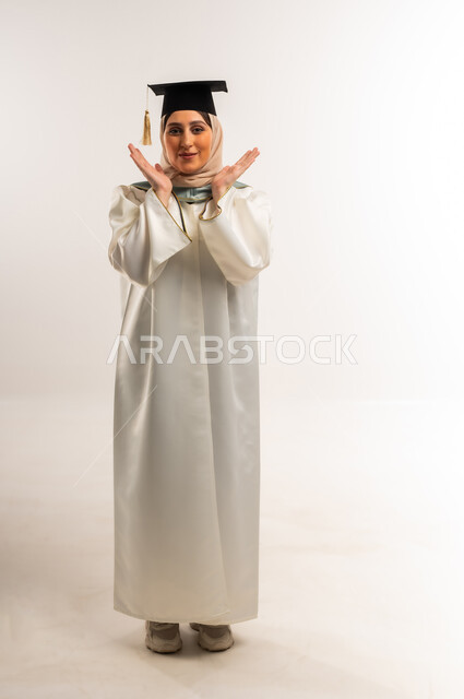 إيماءات الفرح بالوصول لحلم النجاح، بورتريه لخريجة عربية خليجية عمانية محجبة مبتسمة ترتدي زي التخرج والقبعة ترفع يديها تنظر إلى الكاميرا بتعابير السرور والسعادة، حفلة التخرج من الجامعة, شابة سعودية تحتفل بإنهاء الدراسة الجامعية، الجسم بالطول الكامل، خلفية بيضاء