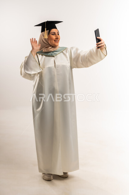 إلقاء التحية والسلام، مشاركة الأهل والأصدقاء فرحة الوصول لحلم النجاح، بورتريه لخريجة عربية خليجية عمانية محجبة ترتدي زي التخرج والقبعة ترفع يدها تقوم بإجراء مكالمة فيديو عبر الهاتف المحمول، شابة سعودية تستخدم جهاز تقني حديث، الجسم بالطول الكامل، خلفية بيضاء