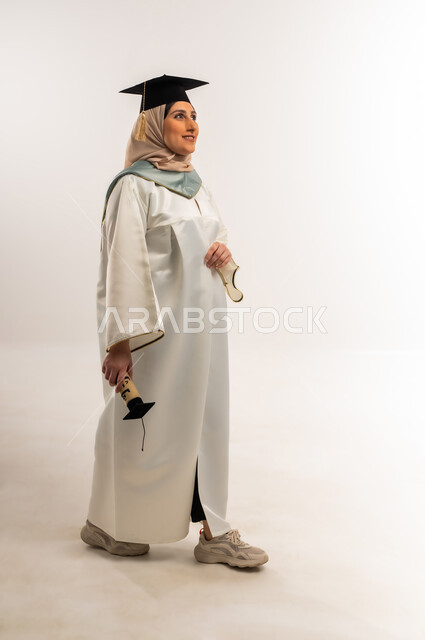 إيماءات الذهاب إلى مكان ما، مناسبة واحتفال جامعي، السعادة بالوصول لحلم النجاح، بورتريه لخريجة عربية خليجية عمانية محجبة ترتدي زي التخرج والقبعة تمسك وثيقة التخرج تنظر إلى شيء ما، شابة سعودية تحتفل بإنهاء الدراسة الجامعية، الجسم بالطول الكامل، خلفية بيضاء