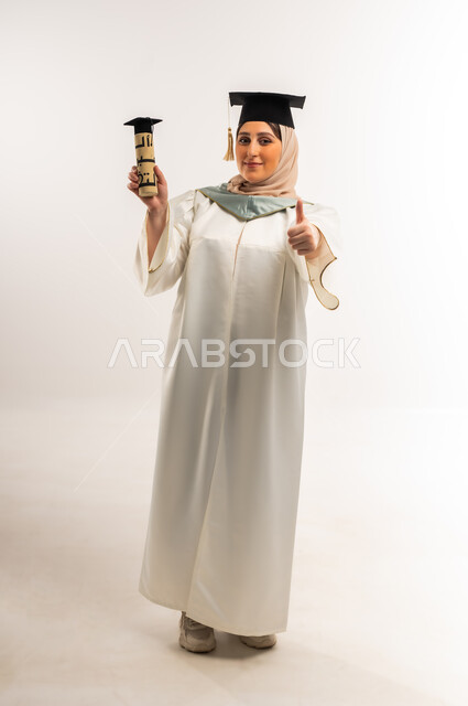 Young Saudi woman celebrating the completion of university studies, a mark of quality and excellence, happiness in reaching the dream of success, portrait of a veiled Gulf Arab Omani graduate wearing a graduation uniform and cap holding a graduation document raising her hand with an expression of joy, university occasion and celebration, full body, white background