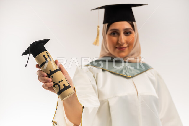 شابة سعودية تحتفل بإنهاء الدراسة الجامعية، إيماءات السعادة بالوصول لحلم النجاح، بورتريه مقرب لخريجة عربية خليجية عمانية محجبة ترتدي زي التخرج والقبعة تمسك بيدها وثيقة التخرج تنظر إلى الكاميرا بتعابير السرور، مناسبة واحتفال جامعي، خلفية بيضاء