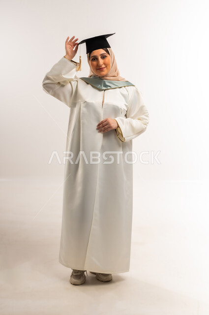 University graduation party, gestures of happiness at reaching the dream of success, portrait of a smiling veiled Gulf Arab Omani graduate wearing graduation uniform and cap raising her hand looking at the camera with expressions of pleasure, Saudi young woman celebrating finishing university studies, full body, white background