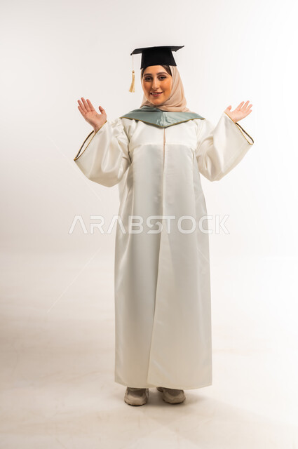 حفلة التخرج من الجامعة, إيماءات السعادة بالوصول لحلم النجاح، بورتريه لخريجة عربية خليجية عمانية محجبة مبتسمة ترتدي زي التخرج والقبعة ترفع يديها تنظر إلى الكاميرا بتعابير السرور، شابة سعودية تحتفل بإنهاء الدراسة الجامعية، الجسم بالطول الكامل، خلفية بيضاء