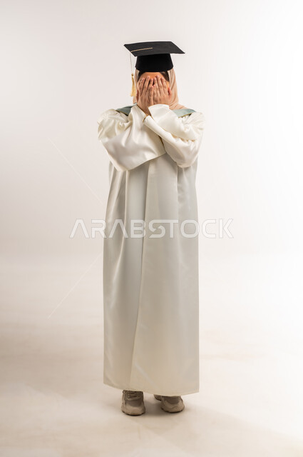 حفلة التخرج من الجامعة، إيماءات البكاء من السعادة بالوصول لحلم النجاح، بورتريه لخريجة عربية خليجية عمانية محجبة مبتسمة ترتدي زي التخرج والقبعة تضع يديها على وجهها بتعابير الخجل، شابة سعودية تحتفل بإنهاء الدراسة الجامعية، الجسم بالطول الكامل، خلفية بيضاء