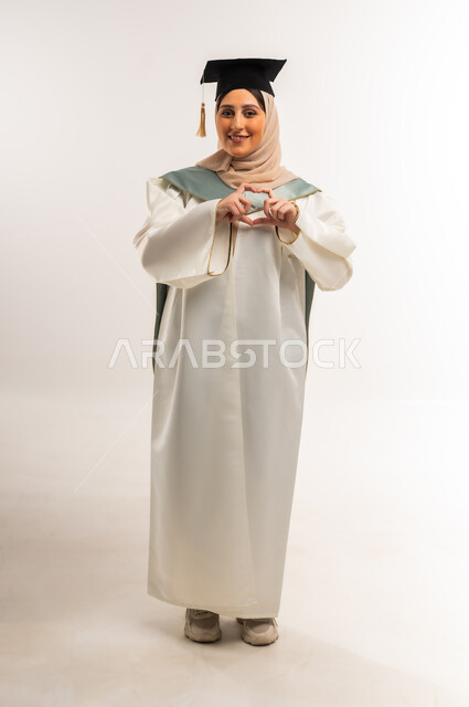 Showing feelings of love and passion, gestures of happiness for reaching the dream of success, portrait of a smiling veiled Gulf Arab Omani graduate wearing graduation uniform and cap, raising her hands in the shape of a heart, looking at the camera with expressions of pleasure, a young Saudi woman celebrating the completion of university studies, full body, white background