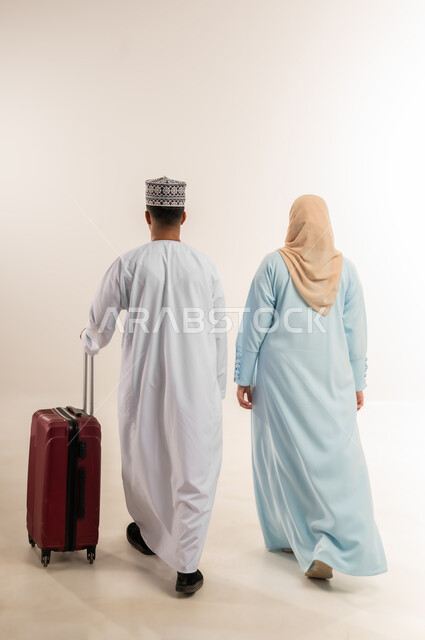 الاستعداد لقضاء الإجازة الصيفية والذهاب في رحلة سفر، زوجان عمانيان يقومان بالمشي إلى مكان ما، بورتريه من الخلف لرجل عربي خليجي عماني يرتدي الدشداشة والكمة يقف مع زوجته يجر حقيبة الأمتعة، الأجواء العائلية المليئة بالحب، الجسم بالطول الكامل، خلفية بيضاء