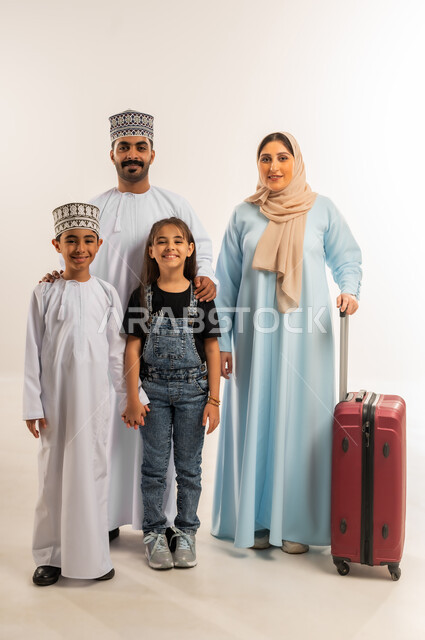 النظر إلى الكاميرا بإيماءات السعادة والسرور، بورتريه لسيدة عربية خليجية عمانية ترتدي العباءة والحجاب تقف مع زوجها وأبنائها تمسك حقيبة الأمتعة، الإستعداد لقضاء الإجازة الصيفية والذهاب في رحلة سفر، الأجواء العائلية المليئة بالحب، الجسم بالطول الكامل، خلفية بيضاء