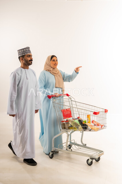 Woman pointing index finger at something, sharing in buying household items, portrait of a gulf arab omani couple pulling a shopping cart, taking advantage of discounts and offers, buying products from a supermarket, walking around a mall and shopping center, full body, white background
