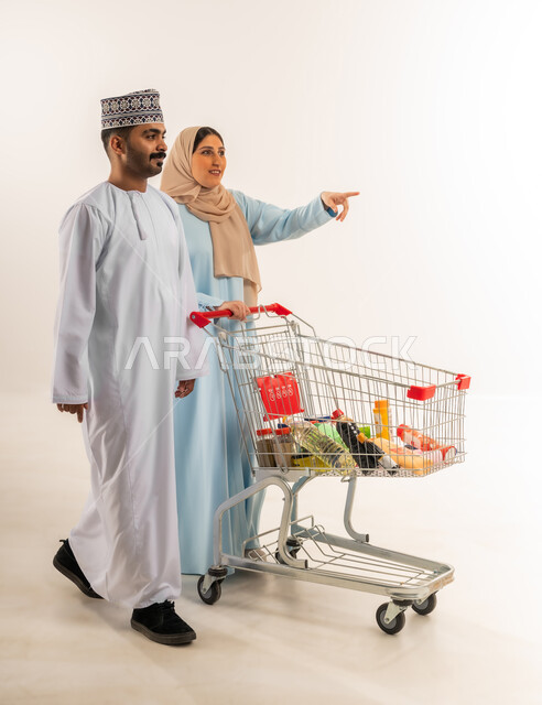 Participating in buying household supplies, portrait of an Arab Gulf Omani couple pulling a shopping cart, taking advantage of discounts and offers, woman pointing index finger at something, buying products from a supermarket, walking around a mall and shopping center, full body, white background