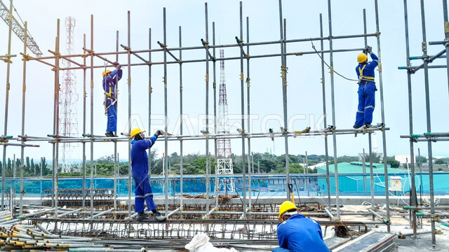 مفهوم الهندسة الانشائية والمعمارية، أعمال البناء وصب الخرسانات ووضع الأساسات الأولية، تنفيذ مخطط الإعمار وتشكيل الهيكل المعدني, مبنى قيد الإنشاء والتشييد في المملكة العربية السعودية, عمال عرب خليجيون سعوديون يرتدون سترة وخوذة الحماية يقومون بوضع السقالات