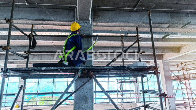 أعمال البناء وصب الخرسانات ووضع الأساسات الأولية، تنفيذ مخطط الإعمار وتشكيل الهيكل المعدني, مبنى قيد الإنشاء والتشييد في المملكة العربية السعودية, عامل عربي خليجي سعودي يرتدي سترة وخوذة الحماية يقوم بفحص السقالة, مفهوم الهندسة الانشائية والمعمارية