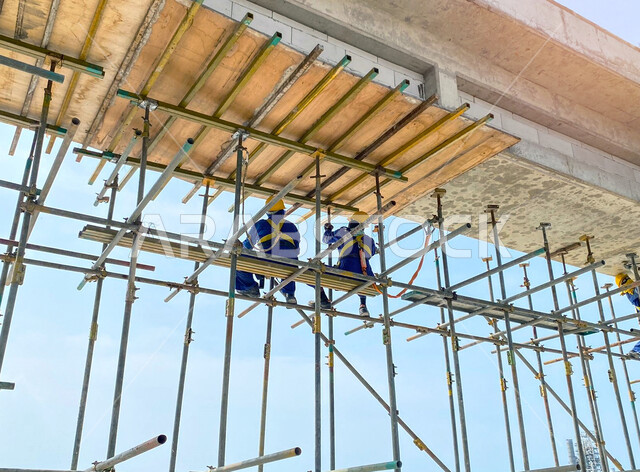 مفهوم الهندسة الانشائية والمعمارية, أعمال البناء وصب الخرسانات ووضع الأساسات الأولية، تنفيذ مخطط الإعمار وتشكيل الهيكل المعدني, صورة من الخلف لعاملين عربيين خليجيين سعوديين يرتديان سترة وخوذة الحماية يقومان بفحص السقالات