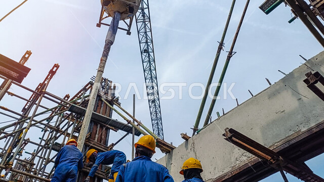 صب الخرسانة باستخدام الآلات والمعدات الثقيلة الضخمة, مفهوم الهندسة الانشائية والمعمارية, أعمال البناء ووضع الأساسات الأولية، تنفيذ مخطط الإعمار وتشكيل الهيكل المعدني, عمال عرب خليجيون سعوديون يرتدون سترة وخوذة الحماية يقفون في موقع العمل