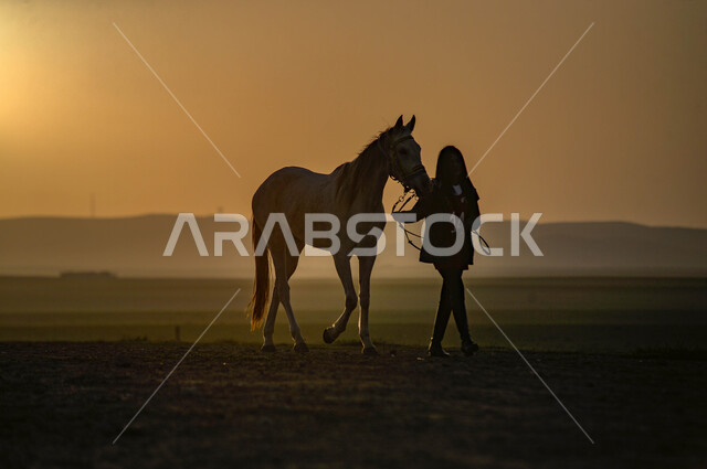 Equestrianism and horse riding hobby, fun recreational activity, interest and passion for horse riding, Saudi Arabian people's attachment to horses, silhouette of a Saudi Arabian Gulf girl wearing casual clothes standing next to brown horses in the desert at sunset, Salulit