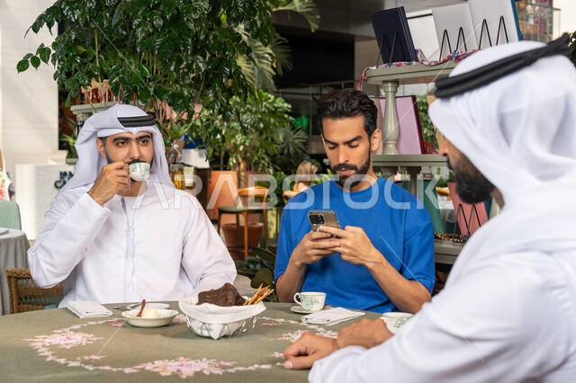 Holiday entertainment and fun, two Emirati Gulf Arab men wearing white kandura and ghutra sitting with their friend in a cafe with gestures of happiness and pleasure, sharing hot drinks, coffee and sweets, using a mobile phone, having a good time outside with friends