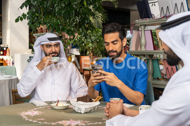 Using mobile phone, having fun outside with friends, having fun and entertainment on vacation, two Emirati Gulf Arab men wearing white kandura and ghutra sitting with their friend in a cafe with gestures of happiness and pleasure, sharing hot drinks, coffee and sweets