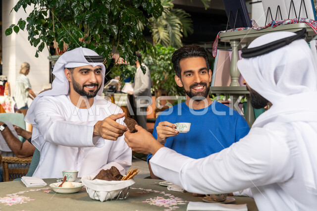 التسلية والترفيه في العطلة، رجلان عربيان خليجان اماراتيان يرتديان الكندورة والغترة البيضاء يجلسان مع صديقهم في المقهى بايماءات السعادة والسرور، مشاركة المشروبات الساخنة والقهوة والحلويات، تبادل الاحاديث بين الأصدقاء، قضاء وقت ممتع في الخارج