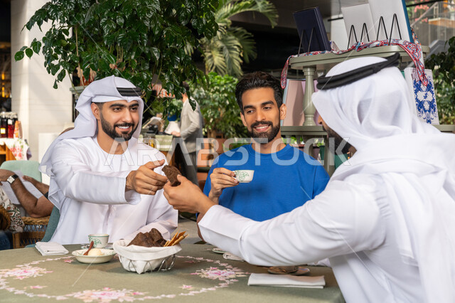 Sharing hot drinks, coffee and sweets, exchanging conversations between friends, spending a good time outside, having fun and entertainment on vacation, two Emirati Gulf Arab men wearing white kandura and ghutra sitting with their friend in the cafe with gestures of happiness and pleasure