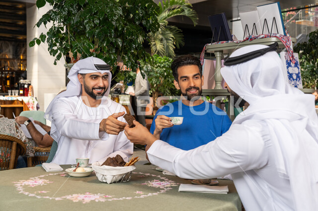 قضاء وقت ممتع في الخارج، التسلية والترفيه في العطلة، رجلان عربيان خليجان اماراتيان يرتديان الكندورة والغترة البيضاء يجلسان مع صديقهم في المقهى بايماءات السعادة والسرور، مشاركة المشروبات الساخنة والقهوة والحلويات، تبادل الاحاديث بين الأصدقاء