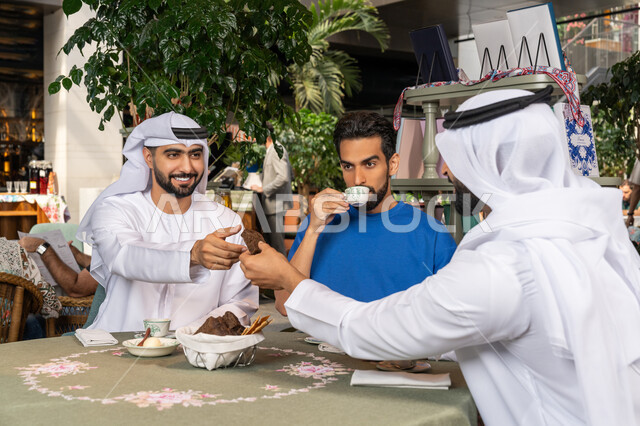 التسلية والترفيه في العطلة، رجلان عربيان خليجان اماراتيان يرتديان الكندورة والغترة البيضاء يجلسان مع صديقهم في المقهى بايماءات السعادة والسرور، مشاركة المشروبات الساخنة والقهوة والحلويات، تبادل الاحاديث بين الأصدقاء، قضاء وقت ممتع في الخارج