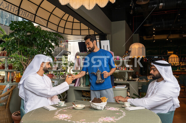 Young man in casual wear serving coffee to his friends, having a good time outside with friends, entertainment and fun on vacation, two arabian gulf emirates men wearing kandura and ghutra sitting in cafe with gestures of happiness and pleasure, sharing hot drinks and sweets