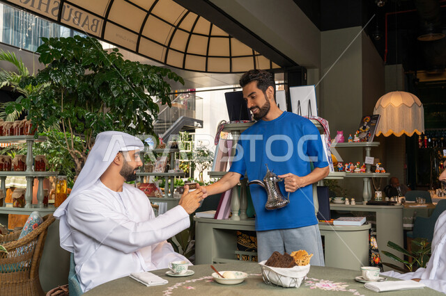 Sharing hot drinks and sweets, young man in casual wear serving coffee to his friends, having a good time outside with friends, entertainment and fun on vacation, two Arab Gulf Emirati men wearing kandura and ghutra sitting in a cafe with gestures of happiness and pleasure