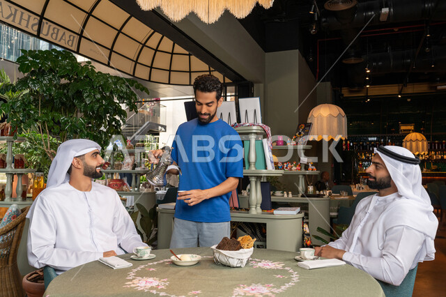 Young man in casual wear serving coffee to his friends, having a good time outside with friends, entertainment and fun on vacation, two arabian gulf emirates men wearing kandura and ghutra sitting in cafe with gestures of happiness and pleasure, sharing hot drinks and sweets