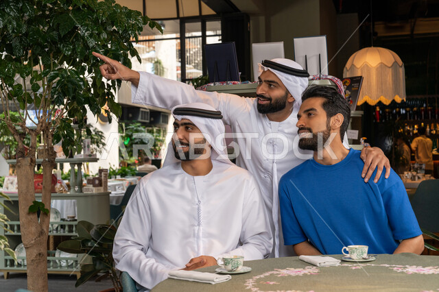 Pointing with the index finger at something, looking at a place with gestures of happiness and pleasure, building successful social relationships between friends, spending a fun and entertaining time with friends, a close-up photo of two Emirati Gulf Arab men wearing kandura and ghutra sitting with their friend in a cafe