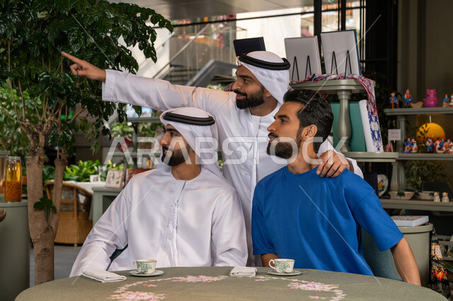 النظر الى مكان ما بايماءات السعادة والسرور،  بناء علاقات اجتماعية ناجحة بين الأصحاب، الاشارة باصبع السبابة إلى شيء ما، قضاء وقت ممتع ومسلي مع الأصدقاء، صورة مقربة لرجلين عربيين خليجيين اماراتيين يرتديان الكندورة والغترة يجلسان مع صديقهم في المقهى