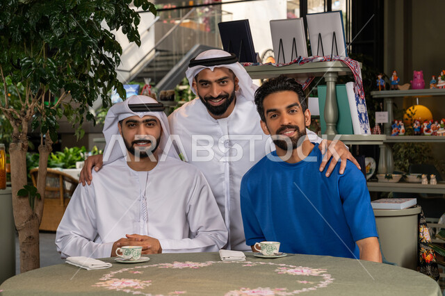النظر الى الكاميرا بايماءات السعادة والسرور،  بناء علاقات اجتماعية ناجحة بين الأصحاب، التقاط  صورة تذكارية وتوثيق اللحظات السعيدة، قضاء وقت ممتع ومسلي مع الأصدقاء، صورة مقربة لرجلين عربيين خليجيين اماراتيين يرتديان الكندورة والغترة يجلسان مع صديقهم في المقهى