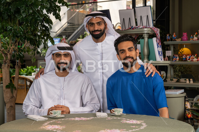 التقاط  صورة تذكارية وتوثيق اللحظات السعيدة، النظر الى الكاميرا بايماءات السعادة والسرور،  بناء علاقات اجتماعية ناجحة بين الأصحاب، قضاء وقت ممتع ومسلي مع الأصدقاء، صورة مقربة لرجلين عربيين خليجيين اماراتيين يرتديان الكندورة والغترة يجلسان مع صديقهم في المقهى